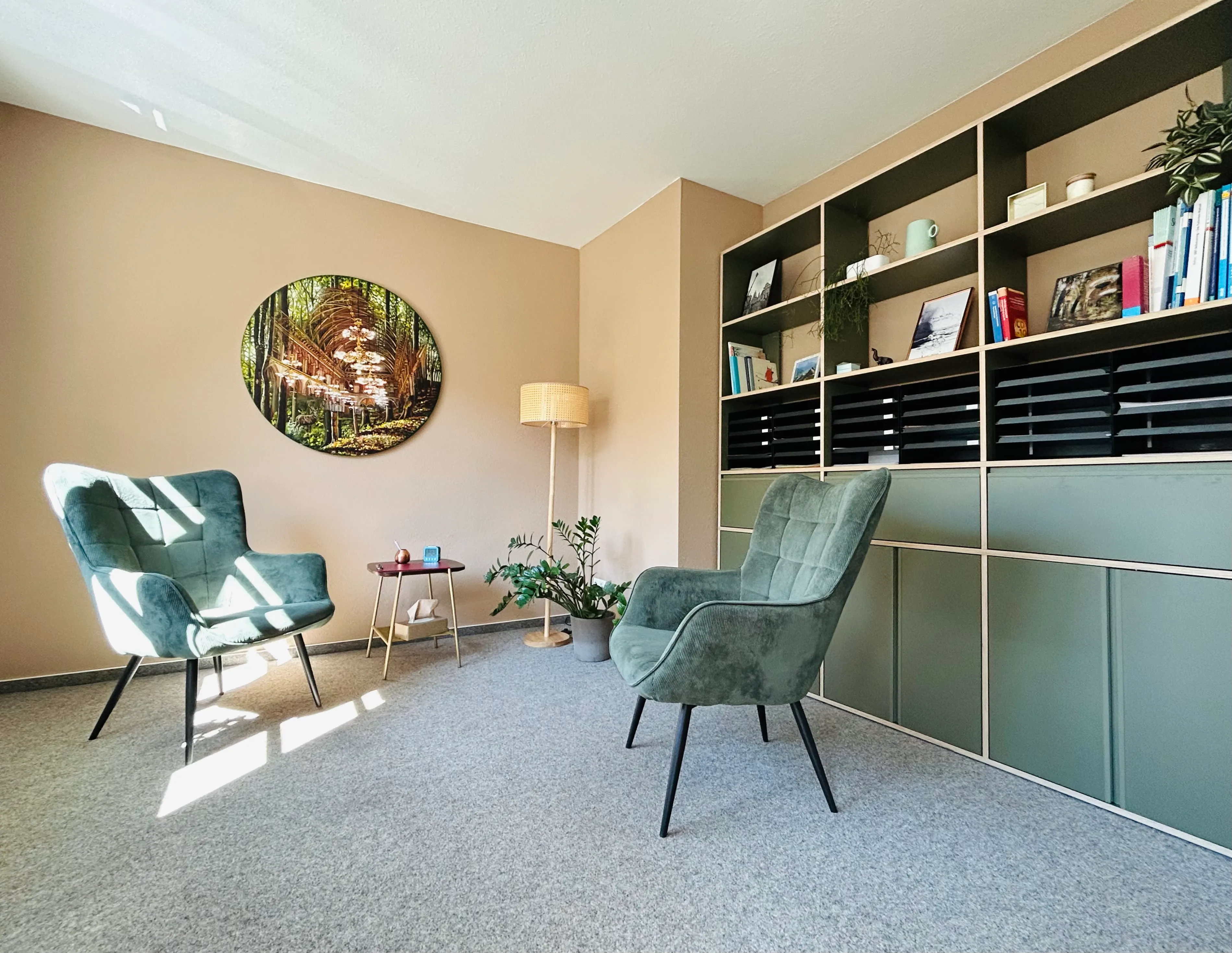 Heller, modern eingerichteter Raum mit zwei grünen Polstersesseln, einem kleinen Beistelltisch und einer Stehlampe. An der Wand hängt ein rundes Bild mit einem Waldmotiv. Rechts befindet sich ein großes, offenes Regal mit Büchern, Dekoration und Pflanzen. Durch die Fenster fällt helles Sonnenlicht auf den Teppichboden.
